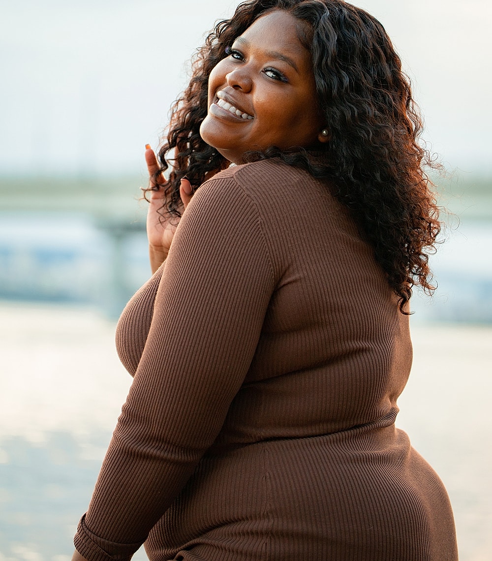 Woman smiling outdoors near water in brown outfit.